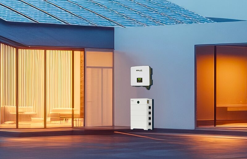 A SolaX wall-mounted inverter and stacked battery unit installed on the exterior wall of a modern house with solar panels on the roof