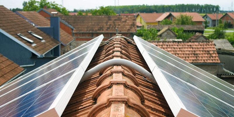Solar panels installed on both sides of a tiled ridge roof with cabling running over the ridge, overlooking a village of tiled rooftops