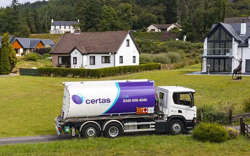 A Certas Energy branded fuel oil tanker lorry driving along a country lane past houses.