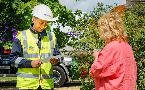 A Certas Energy heating oil delivery driver in hi-vis vest and hard hat talking to a woman in her garden with a tanker behind