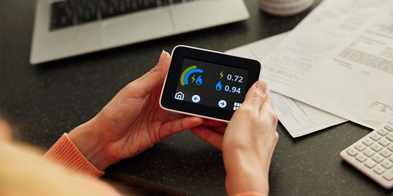 A person holding a smart energy display showing electricity and gas readings, with a laptop, calculator and paperwork on a desk