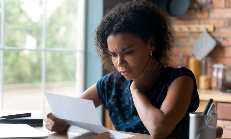 A concerned woman with curly hair reading a letter at a wooden table in a sunlit kitchen