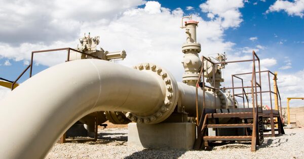 A large industrial gas pipeline with valves and metal access stairs at an outdoor processing site under a blue sky