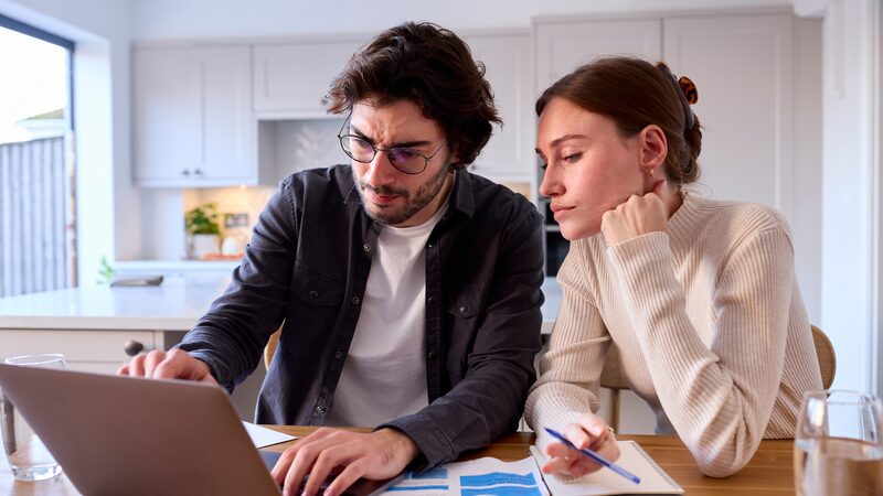 A couple sitting at a kitchen table looking at a laptop with energy bills and a notebook