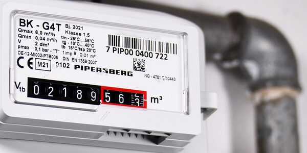 A close-up of a white gas meter with a mechanical dial reading, mounted next to pipework