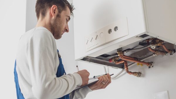An engineer in blue overalls inspecting a wall-mounted white gas boiler and taking notes on a clipboard