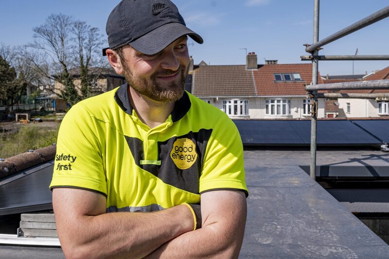 A Good Energy installer in a hi-vis polo shirt and cap standing on a rooftop next to solar panels