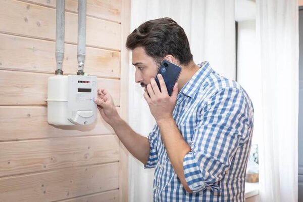 A man on a mobile phone reading a wall-mounted gas meter in a home