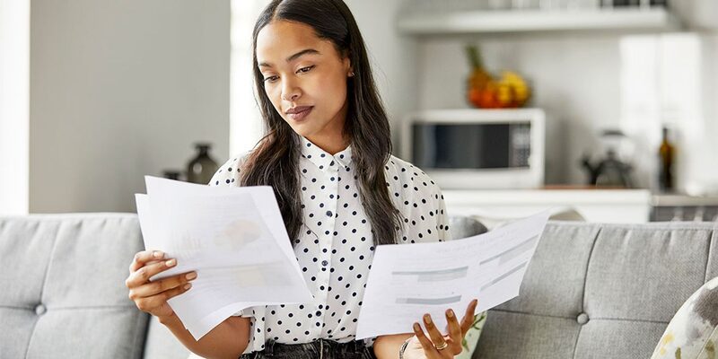 A woman sitting on a sofa at home comparing two printed energy bills or documents