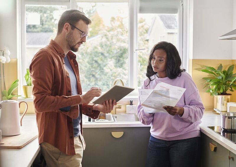 A couple in a kitchen reviewing a paper energy bill together while one holds a tablet