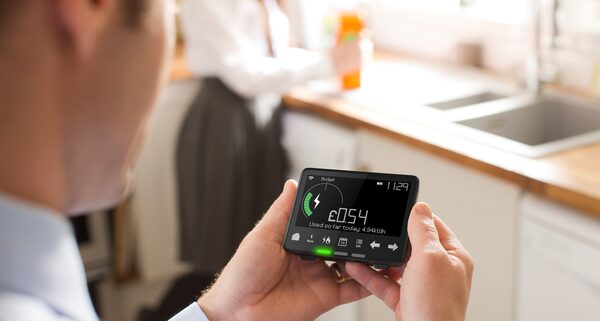 A man holding a smart meter in-home display showing £0.54 of energy used so far today, in a kitchen