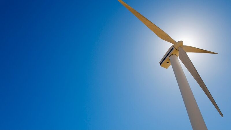 A wind turbine photographed from below against a bright blue sky with the sun behind its blades