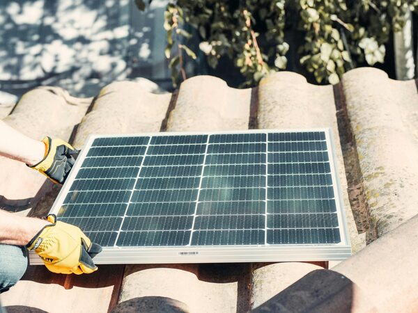 A gloved installer lifting a solar panel onto a terracotta tiled roof