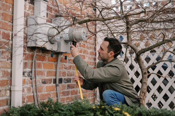 A man plugging a yellow cable into an electricity meter mounted on a brick house wall
