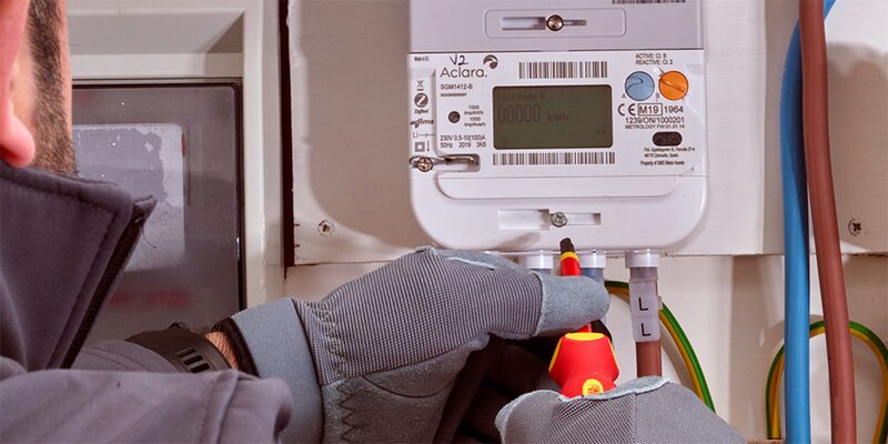 A technician in gloves using a screwdriver to install an Aclara electricity smart meter on a wall