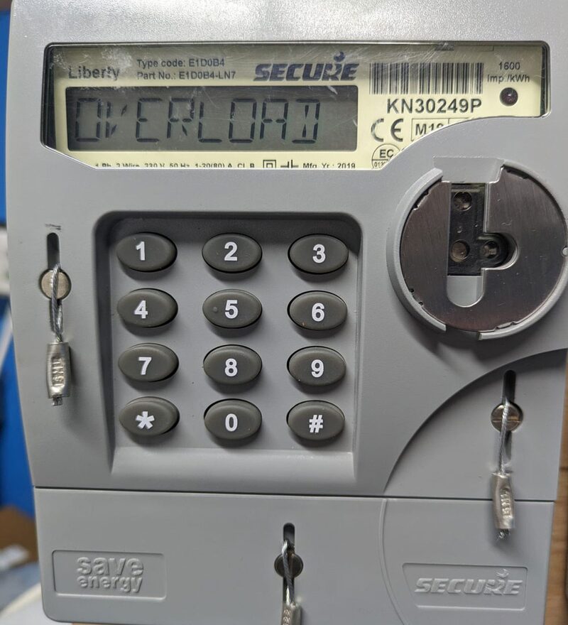 A close-up of a Secure Liberty prepayment electricity meter displaying 'OVERLOAD' with a keypad and key slots
