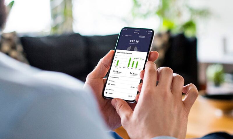 A person holding a smartphone showing an energy app with a £12.18 balance, bar charts and kWh figures, in a living room