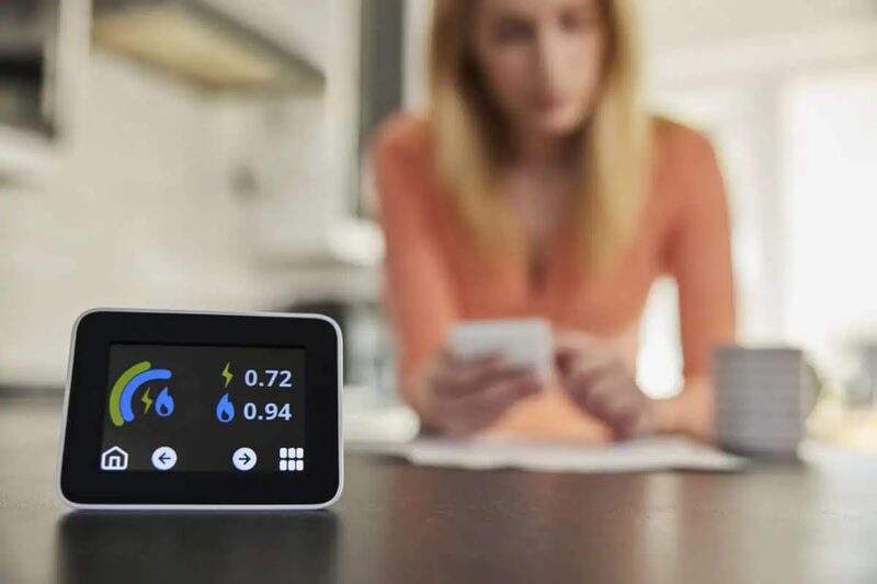 A smart energy monitor display on a kitchen counter showing electricity and gas readings, with a blurred woman reviewing bills behind