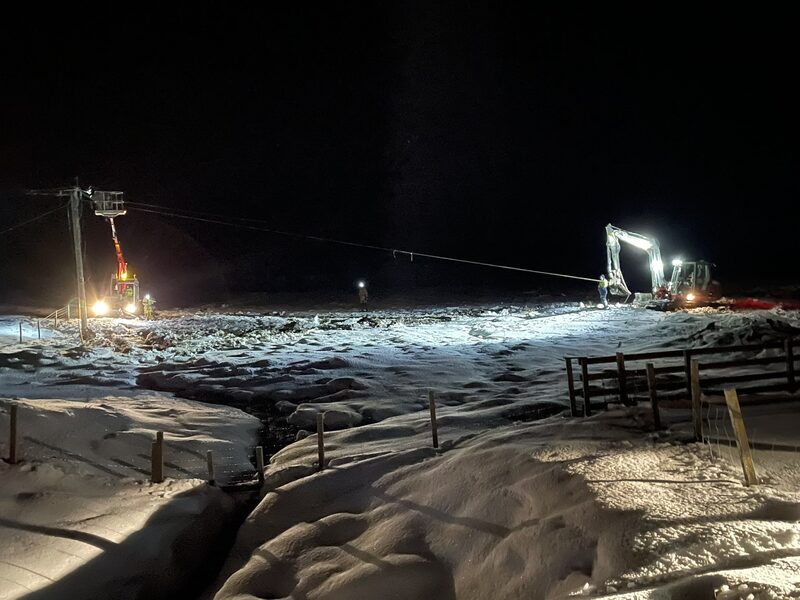 Engineers repairing power lines at night in snowy Shetland countryside, with a cherry picker and excavator working under floodlights