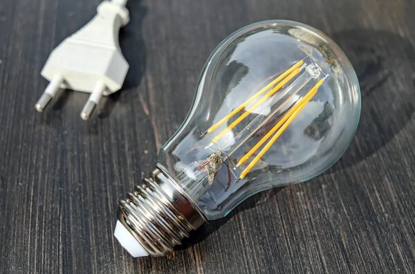 A clear LED filament light bulb lying on a dark wooden surface next to a white two-pin European plug