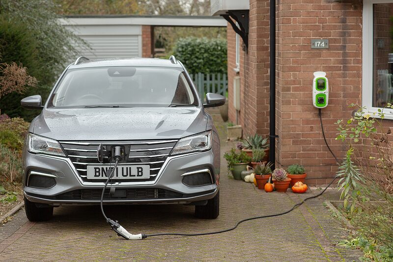 A grey electric car charging on a driveway from a wall-mounted home EV charge point on a brick house