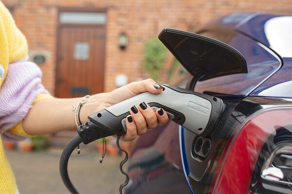A woman plugging a Wallbox EV charging cable into the charging port of a blue car outside a brick house