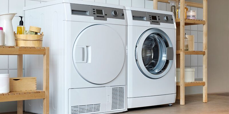 A white tumble dryer and washing machine side by side in a utility room with wooden shelving