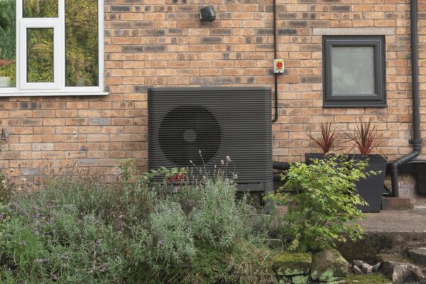 A dark grey air source heat pump unit installed on the exterior brick wall of a house beside a window and garden plants