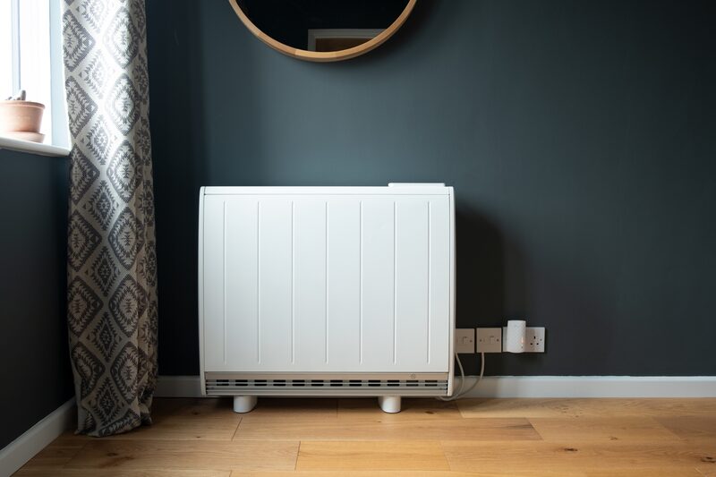 A white electric storage heater installed against a dark green wall in a room with wooden flooring and patterned curtains