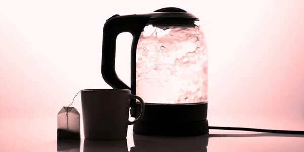 A glass electric kettle boiling beside a mug and a teabag