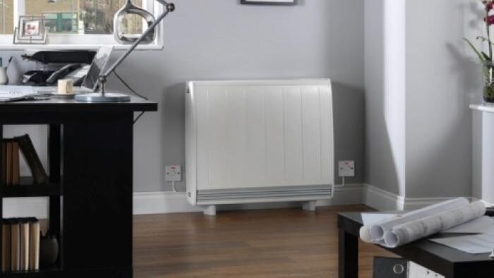 A white electric storage heater installed against the wall of a grey home office with a desk and lamp
