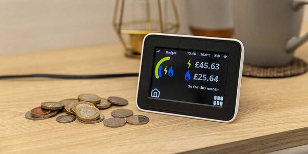 A smart energy monitor on a wooden table showing electricity and gas costs next to a pile of coins