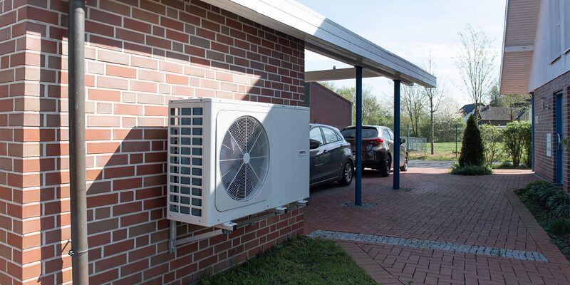 A white air source heat pump unit mounted on the brick wall of a house beside a carport with parked cars