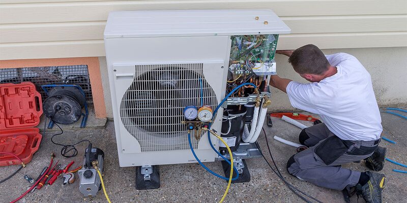 A technician installing an air source heat pump unit outside a house, with tools and pressure gauges around it