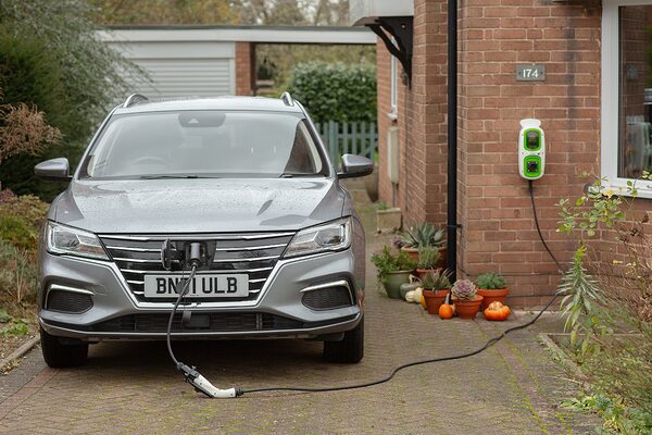A grey electric car charging on a driveway from a wall-mounted home EV charge point on a brick house