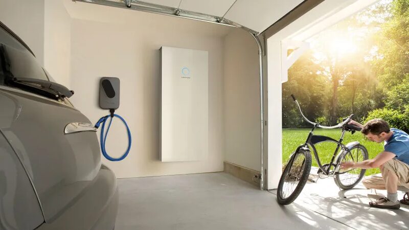A sonnenBatterie home battery and wall-mounted EV charger installed in a garage, with a car parked beside them and a man repairing a bike in the open doorway