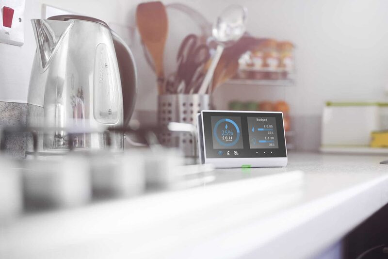 A smart meter display showing energy use and budget sitting on a kitchen counter beside a kettle