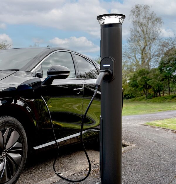 A black electric car charging from a tall dark grey Ratio EV charge post with a light on top, outdoors on a driveway