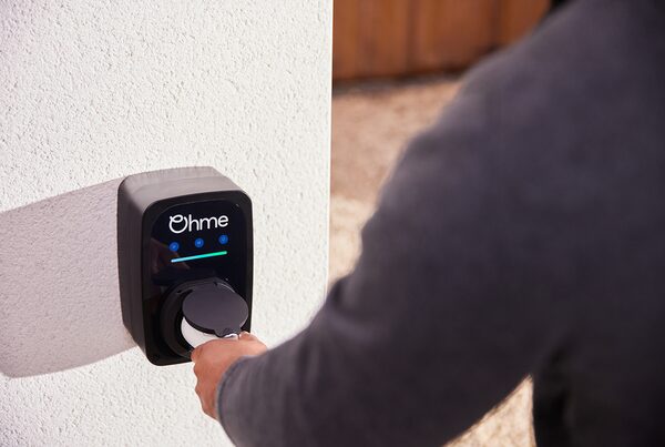 A person plugging a charging cable into a black Ohme wall-mounted EV charger on a white exterior wall