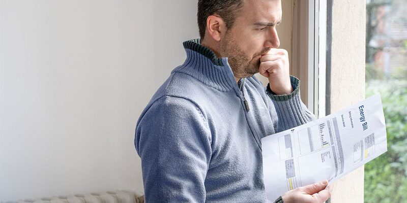 A worried man in a jumper reading an energy bill document beside a window