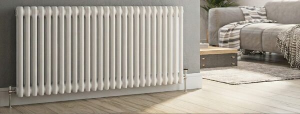 A white column radiator installed on a grey wall in a living room with a sofa in the background
