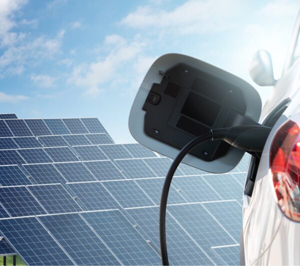 An electric car being charged with a cable plugged into its open charge port, in front of a large solar panel array under a blue sky