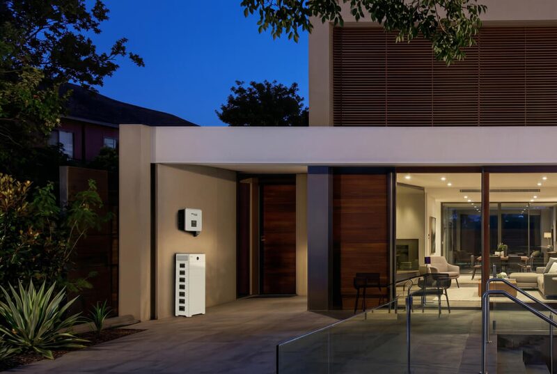 A wall-mounted solar inverter and a tall battery storage unit installed on the exterior wall of a modern house at dusk