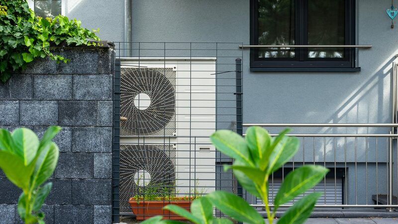 An air source heat pump unit with two fans installed outside a grey house behind a metal guard fence