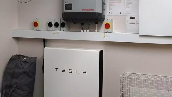 A Tesla Powerwall home battery installed on a wall beneath a Fronius Symo inverter and electrical isolators in a utility room