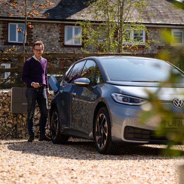 A man charging a grey Volkswagen ID.3 electric car from a wall-mounted EV charger on a gravel driveway outside a brick house