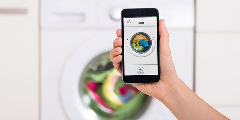 A hand holding a smartphone showing a washing machine control app in front of a blurred washing machine