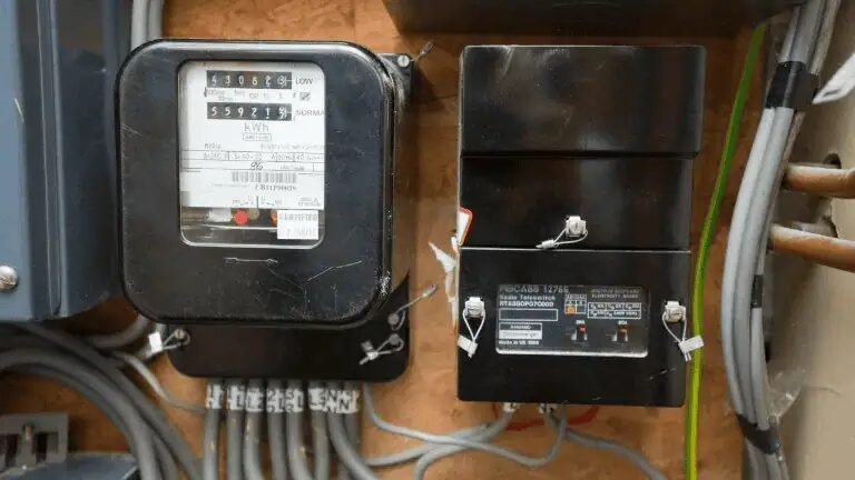 A traditional electricity meter mounted on a wall beside a separate grey teleswitch box, with wiring from the teleswitch running to the meter and onward to a storage heater and an immersion-heated hot water cylinder, showing it switches both the tariff rate and the heating circuits.