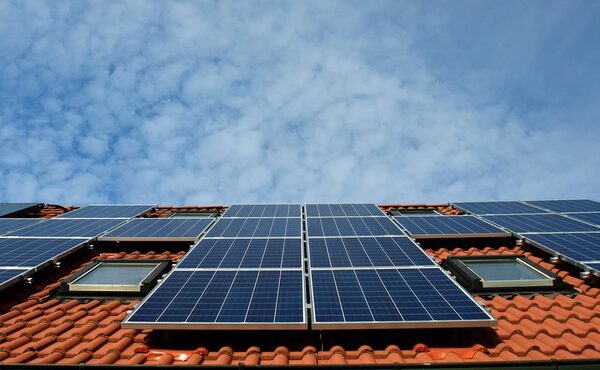 Solar panels installed on a red-tiled house roof with roof windows, under a blue cloudy sky