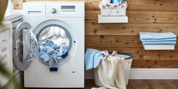 A white front-loading washing machine with its door open and laundry inside, next to a laundry basket in a wood-panelled utility room
